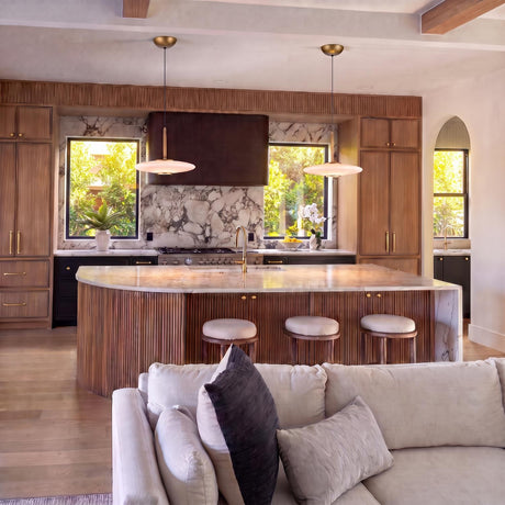 A modern kitchen with two Callan LED Pendants in aged brass and opal glass suspended over a fluted wood island with marble countertops and wooden ceiling beams.
