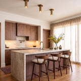 A contemporary kitchen with three Wilber 2 inch Cylinder LED Flush Mounts in aged brass installed along the ceiling over a marble waterfall island and walnut cabinetry.