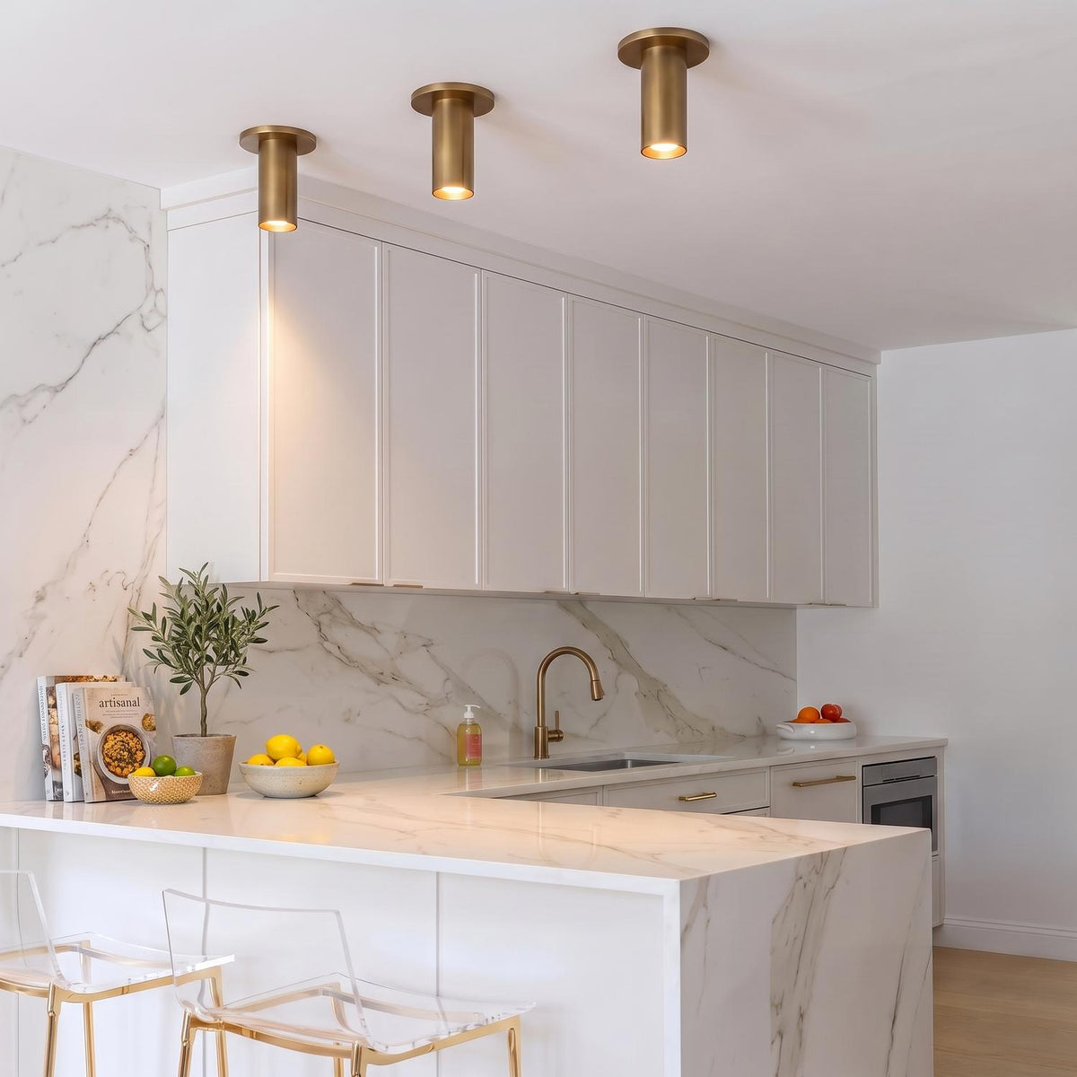 A contemporary kitchen features three Wilber 2" Cylinder LED Flush Mount, Aged Brass fixtures along the ceiling above a marble waterfall counter and white cabinets.