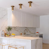 A contemporary kitchen features three Wilber 2" Cylinder LED Flush Mount, Aged Brass fixtures along the ceiling above a marble waterfall counter and white cabinets.