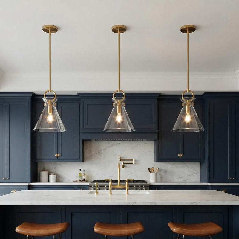 A transitional kitchen features three Aris Small Pendant 11", Aged Brass and Clear Glass fixtures suspended over the white marble island and navy blue cabinetry.