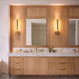 A contemporary bathroom vanity with two Linden LED Wall Sconce 26", Aged Brass fixtures mounted on light wood paneling above a marble countertop.