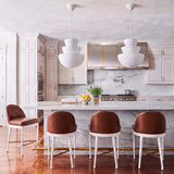 A transitional kitchen with two Verity Pendant 18 inch Textured White pendants over a white marble waterfall island with brown leather bar stools and a brass-trimmed hood.