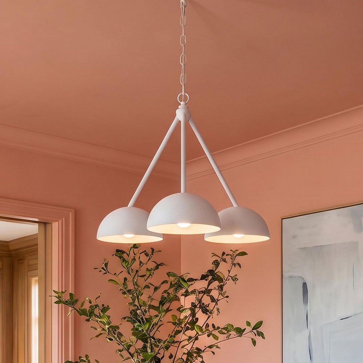 A contemporary living room with peach walls and the Veda 3 Light Chandelier 26", Textured White suspended from a standard ceiling above a leafy green plant.