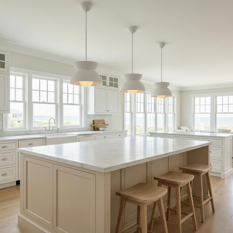 A bright coastal kitchen features three Varo Pendant 15", Textured White fixtures over the white marble island alongside wooden bar stools and cabinetry overlooking the ocean.