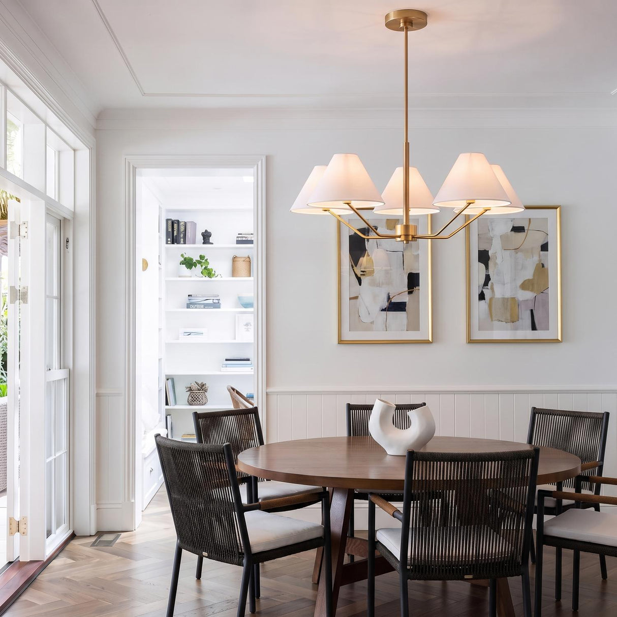 A transitional dining room with the Wallace 5 Light Chandelier in satin brass with white linen shades over a round wood table and woven chairs.