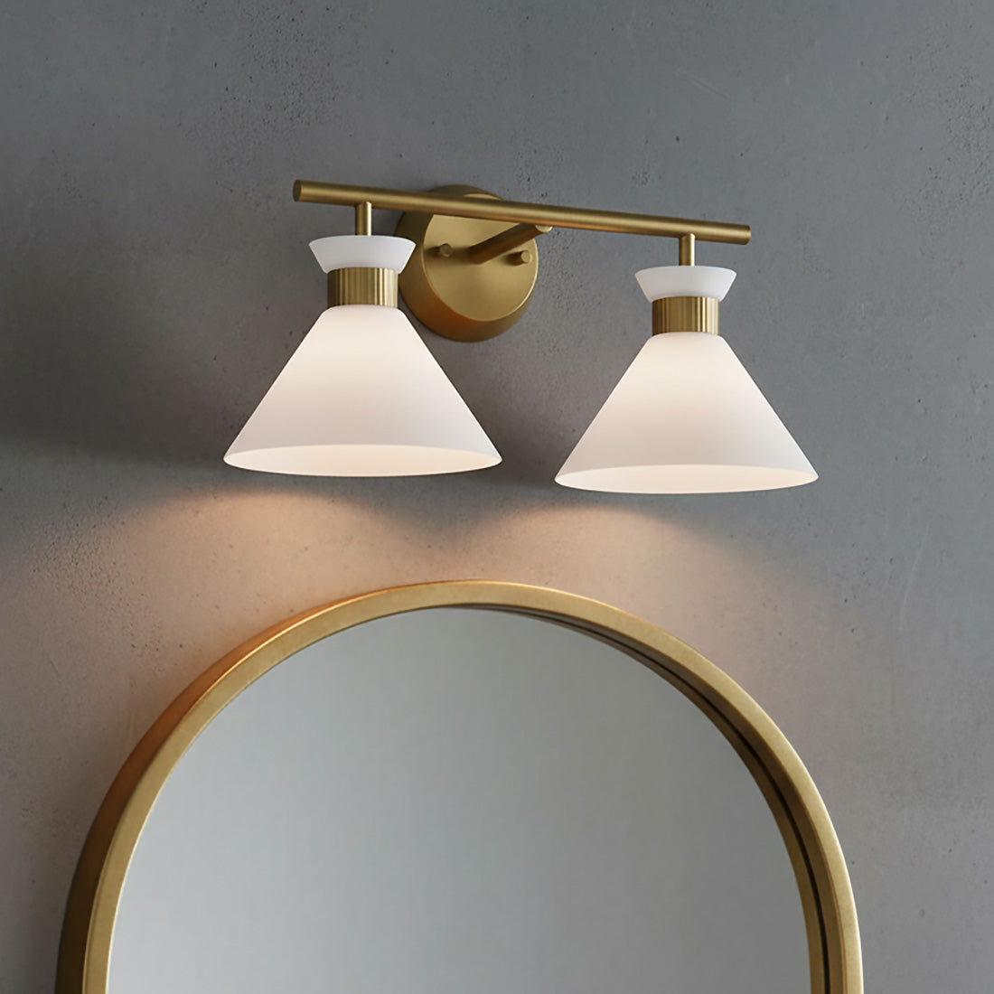 Issa 2 Light Vanity - Two cone-shaped white frosted glass shades with satin brass hardware mounted on a gray wall above a round gold-framed mirror