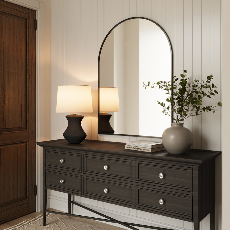 Laken 26" tall Table Lamp, in Textured Black finish, on a dark wood console table with an arched mirror, art books, and a vase with branches.