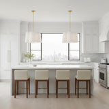 Odelia Large Pendant 28, White and Brass - Two white lamps with brass accents hang over a white kitchen island with four cream-colored bar stools and a cityscape visible through a large window.