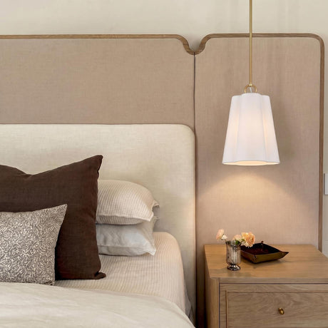 A contemporary bedroom with a white ceramic and brushed brass Odelia Small Pendant hanging over a light wood nightstand beside a large beige fabric headboard.