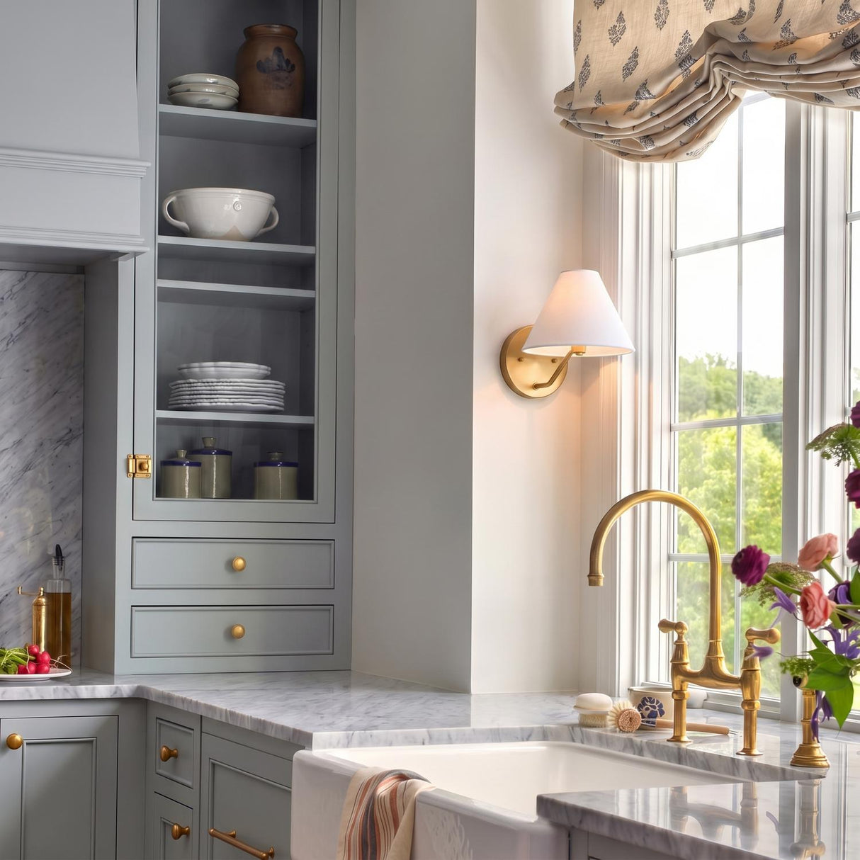 A traditional kitchen featuring the Wallace Wall Sconce 8", Satin Brass with a white linen shade mounted beside a window over a marble farmhouse sink.