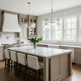 A transitional kitchen featuring two Winslow Chandelier 24", Modern Brass fixtures with textured glass over a white marble island surrounded by taupe cabinetry and upholstered barstools.