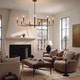 A modern farmhouse living room with an antique iron Thayer 9 Light Chandelier 48" centered over a tan ottoman coffee table beside a white plaster fireplace.