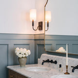 A transitional bathroom with an antique iron wall sconce featuring a linen shade beside a gold framed mirror above a white marble countertop and gray wainscoting.