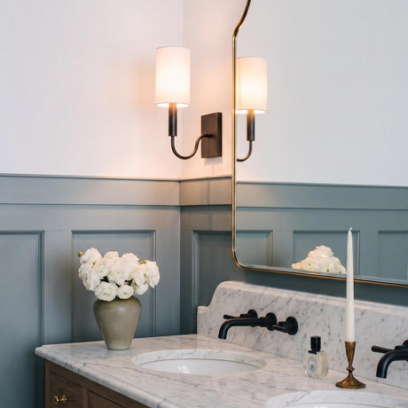A transitional bathroom with an antique iron wall sconce featuring a linen shade beside a gold framed mirror above a white marble countertop and gray wainscoting.
