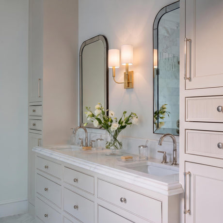 A transitional bathroom featuring a Thayer Double Wall Sconce 14", Modern Brass with linen shades placed between two arched mirrors over a white marble vanity.