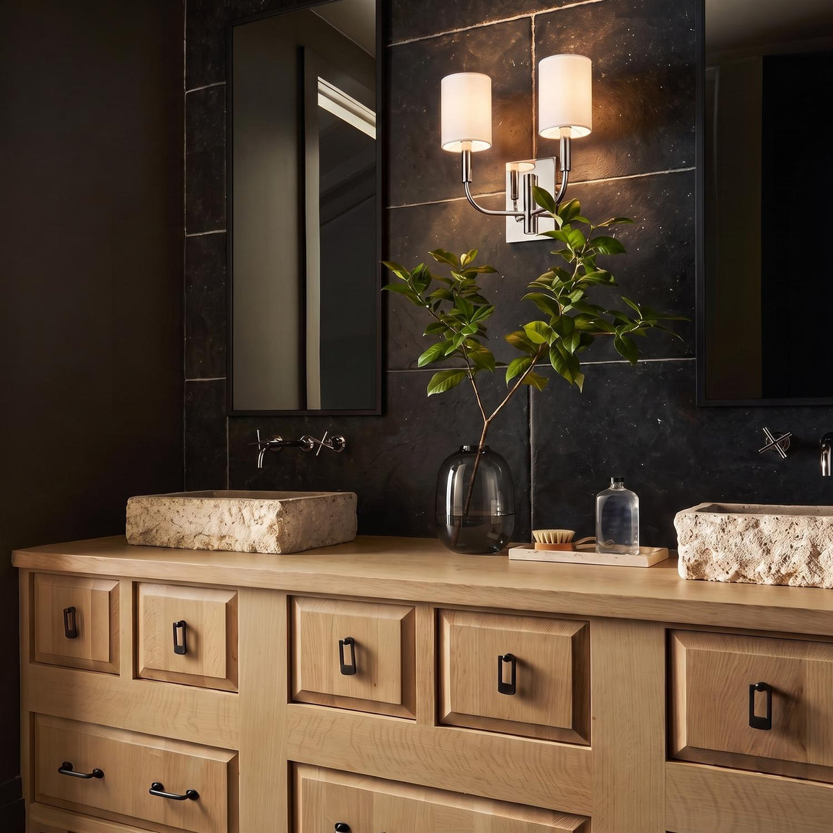A modern bathroom featuring a Thayer Double Wall Sconce 14", Polished Nickel with linen shades mounted on a dark slate wall above two stone vessel sinks.