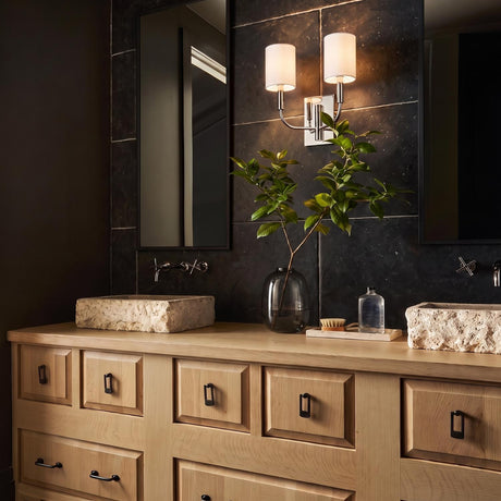 A modern bathroom featuring a Thayer Double Wall Sconce 14", Polished Nickel with linen shades mounted on a dark slate wall above two stone vessel sinks.
