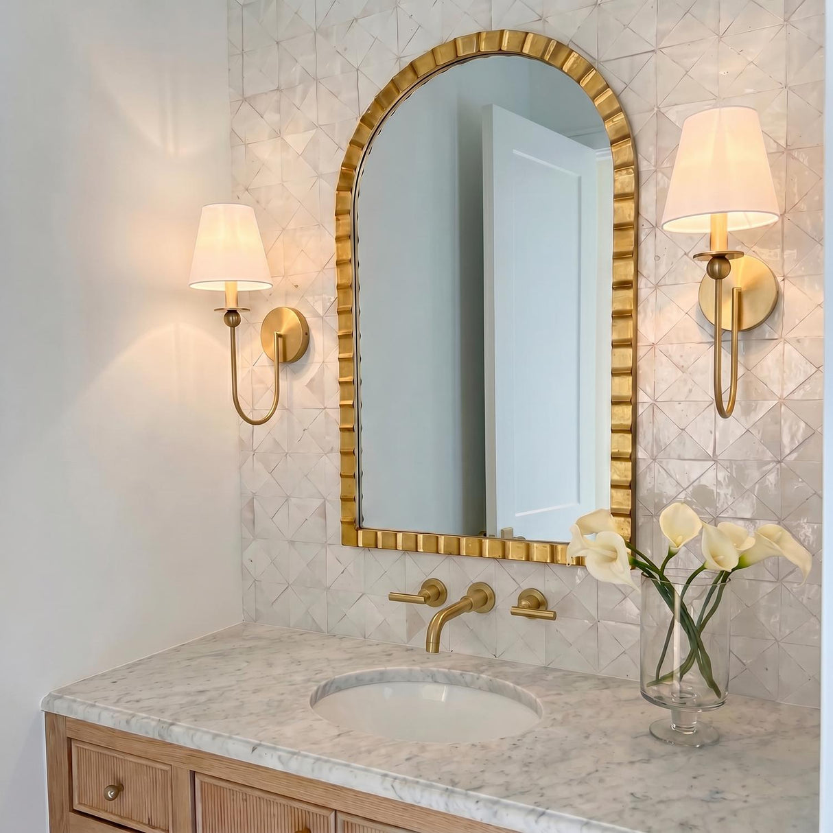 A transitional bathroom with two satin brass wall sconces featuring linen shades flanking a gold arched mirror above a marble countertop and fluted wood vanity.