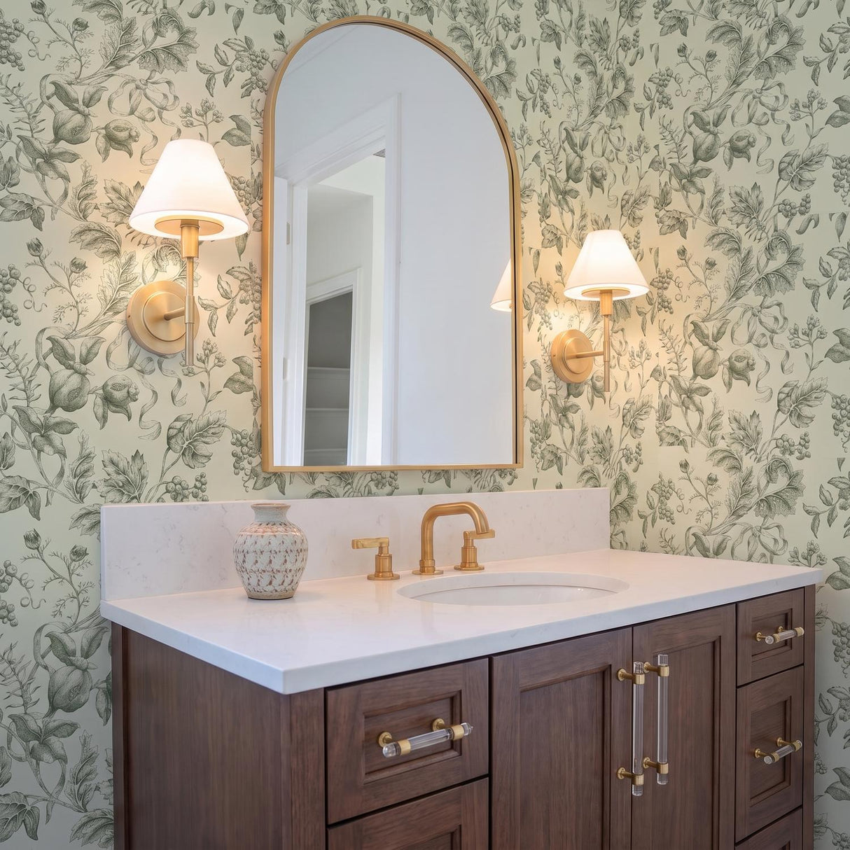 A traditional bathroom featuring two Dean Wall Sconce 14" Satin Brass and Opal Glass fixtures flanking an arched mirror above a white quartz vanity and botanical wallpaper.