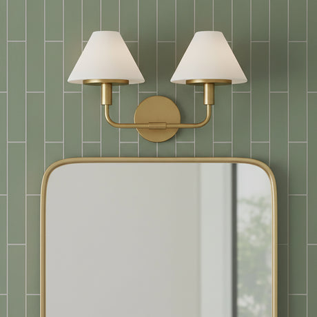 Dean 2 Light Vanity - Two white glass conical shades with satin brass arms and backplate mounted above a rounded corner mirror with a satin brass frame, set against a green subway tile wall.