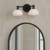 Merrick 2 Light Vanity, Aged Iron - A black two-light vanity fixture with globe shades is mounted above an arched mirror on a wall with vertical cream tiles.