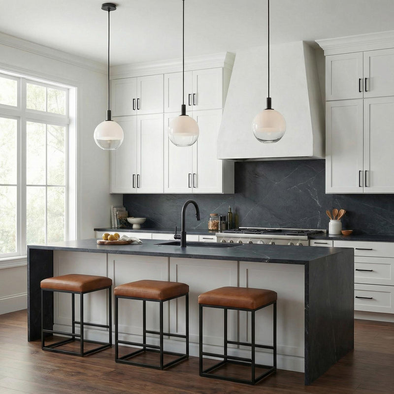 A modern kitchen features three Merrick Pendant 8", Aged Iron fixtures with white globe shades over a dark waterfall island and black marble backsplash with white cabinets.