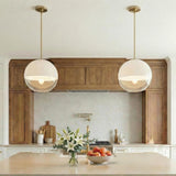 A contemporary kitchen featuring two Merrick Pendant 14", Modern Brass globe pendants with glass shades suspended over a beige island in front of wood cabinetry and backsplash.