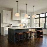 A transitional kitchen features three Merrick Pendant 14", Aged Iron globe pendants with frosted glass over a gray island with white marble and woven bar stools.