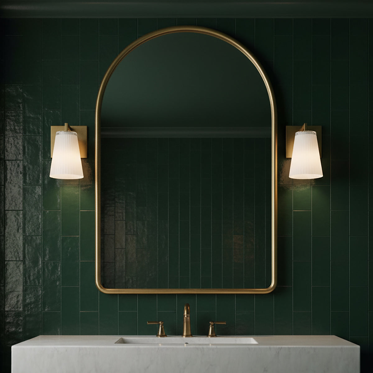 Two Margot 8" tall Wall Sconces, in Satin Brass, shown next to an arched brass vanity mirror against dark green subway tiles.