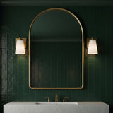 Two Margot 8" tall Wall Sconces, in Satin Brass, shown next to an arched brass vanity mirror against dark green subway tiles.