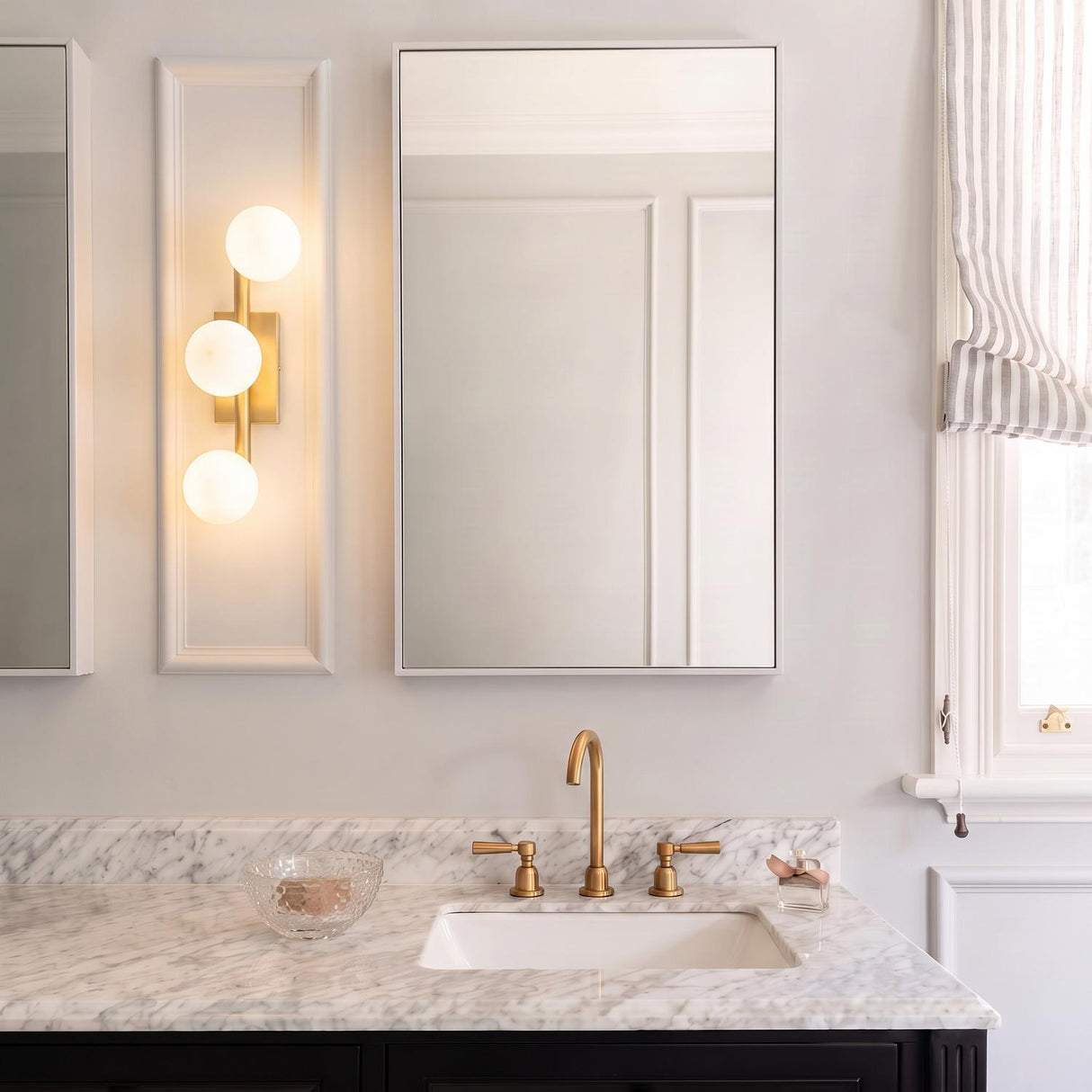 A transitional bathroom with the Veya 3 Light Wall Sconce 16", Modern Brass and Milk Glass mounted beside a mirror over a marble countertop and brass faucet.