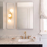 A transitional bathroom with the Veya 3 Light Wall Sconce 16", Modern Brass and Milk Glass mounted beside a mirror over a marble countertop and brass faucet.