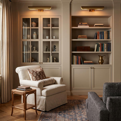 A transitional living room with two Denton Picture Light 18", Modern Brass fixtures mounted above built-in cabinetry beside a cream armchair and rattan side table.
