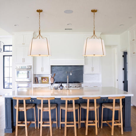 A transitional kitchen featuring two Isleton Large Pendants in aged gilded brass with linen shades over a white quartz island and dark navy blue cabinetry.