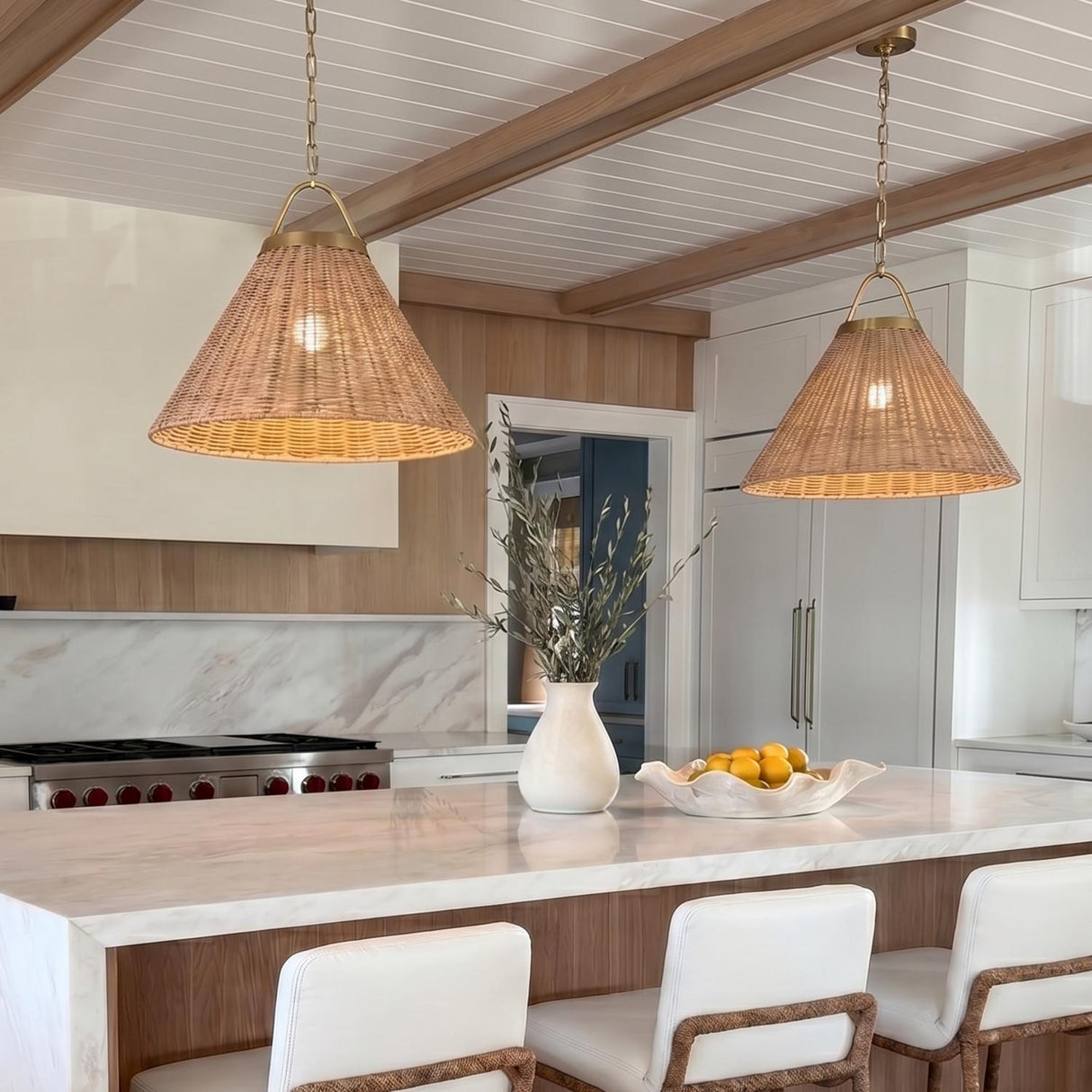 A coastal kitchen with two Paloma Pendant 22" Modern Brass and Rattan pendants over a white marble waterfall island with white cabinetry and a beamed ceiling.