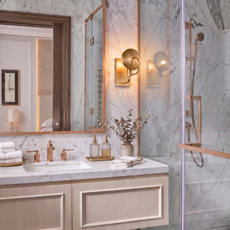 A modern bathroom featuring the Arlen Wall Sconce 10", Modern Brass mounted on white marble wall tile beside a wood-framed mirror and vanity.
