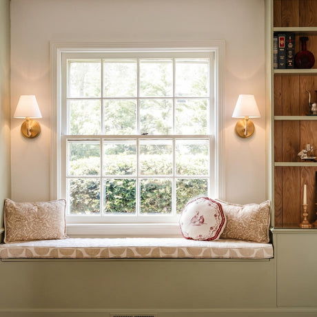 A traditional living room featuring two modern brass and rattan wall sconces with linen shades flanking a window seat and built-in green shelving.