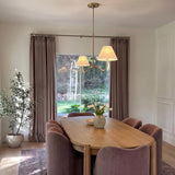 A contemporary dining room with a modern brass linear pendant featuring white linen shades suspended over an oval oak table surrounded by mauve velvet dining chairs.