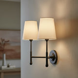 Emerson Double Wall Sconce 14 - A double wall sconce with aged iron finish and cream linen shades is mounted on a textured wall, casting warm light.