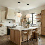 A transitional kitchen features two Emerson Large Pendant 14" Aged Iron globe pendants over a white marble island with natural wood cabinets and woven stools.