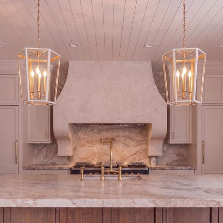 A transitional kitchen features two matte white and brushed brass lantern pendants hanging over a marble island with a matching brass faucet and plaster range hood.