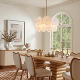 A contemporary dining room featuring the Ophelia Medium Chandelier with modern brass and layered glass discs hanging over a wood pedestal dining table and cream chairs.