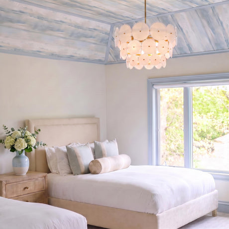 A coastal bedroom featuring the Ophelia Large Chandelier 28", Modern Brass and Layered Glass, suspended from a vaulted blue painted ceiling above a beige upholstered bed.