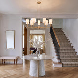 A transitional foyer features the Remington 9 Light Chandelier 36", Satin Brass hanging over a round marble table, with herringbone wood flooring and a carpeted staircase.
