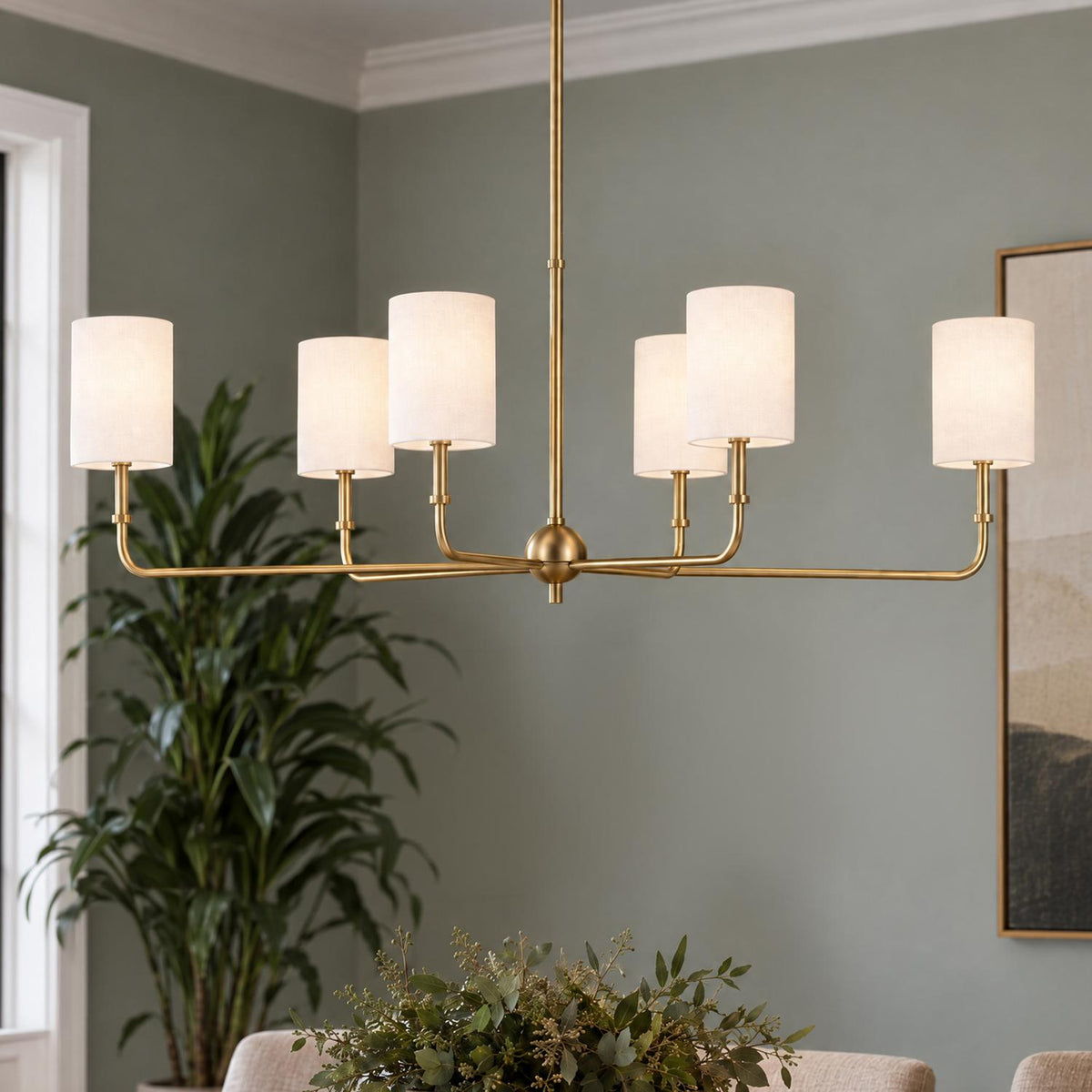 A transitional dining room with the Remington 6 Light Linear Chandelier 44", Satin Brass featuring white fabric shades suspended above a table beside a tall potted plant.