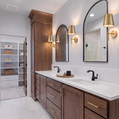 A transitional bathroom with three satin brass wall sconces flanking arched mirrors over a walnut double vanity featuring a white quartz countertop and black faucets.