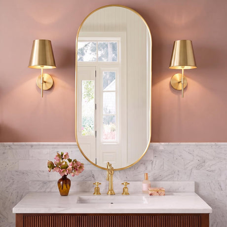 A contemporary bathroom with two satin brass wall sconces flanking a tall gold oval mirror above a white marble backsplash and fluted wood vanity.
