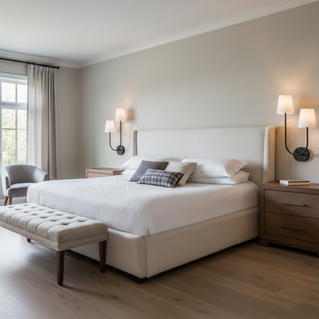 Greta Double Wall Sconce, Black - Two black wall sconces with white shades illuminate a bedroom with a large upholstered bed, a gray armchair, and a wood bench.