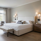 Greta Double Wall Sconce, Black - Two black wall sconces with white shades illuminate a bedroom with a large upholstered bed, a gray armchair, and a wood bench.